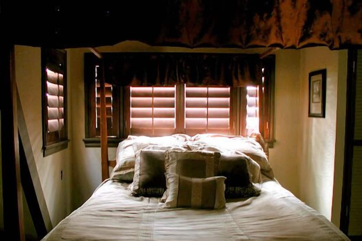 A bed with a headboard, pillows, and a blanket in a room with windows and curtains.