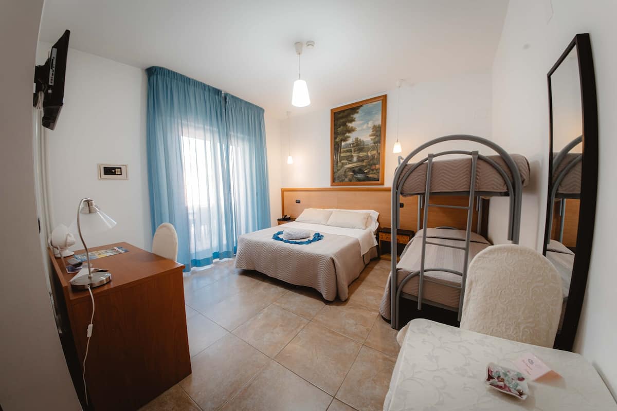 Hotelkamer met een houten bureau, twee bedden, een stapelbed, een bureaulamp, een schilderij aan de muur en een grote spiegel.
