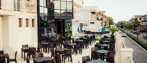 An outdoor cafe with multiple tables and chairs, a modern building with large windows, and a street view in the background.