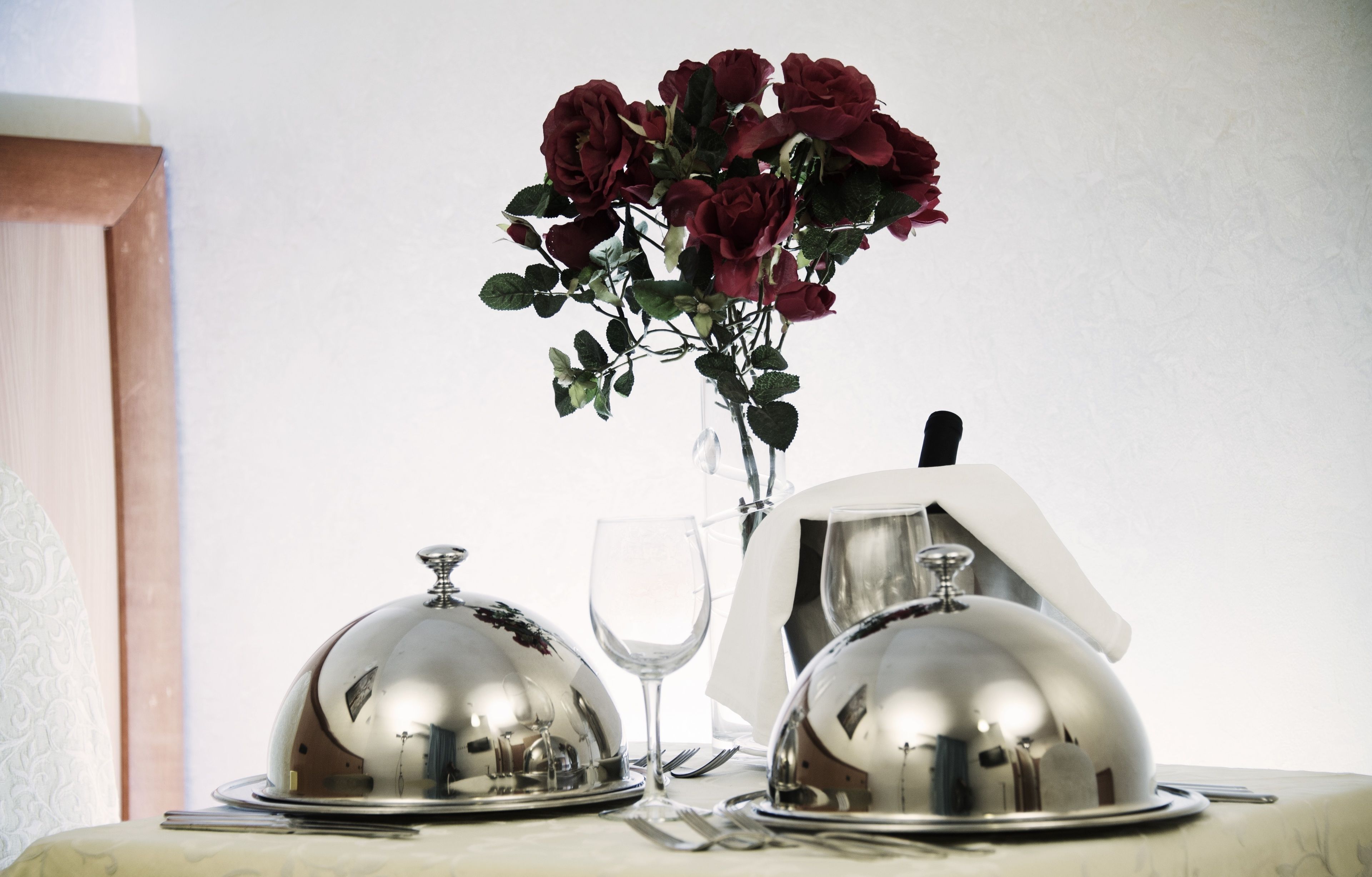 Une table avec deux plats recouverts de cloches en argent, un verre à vin et un bouquet de roses rouges.