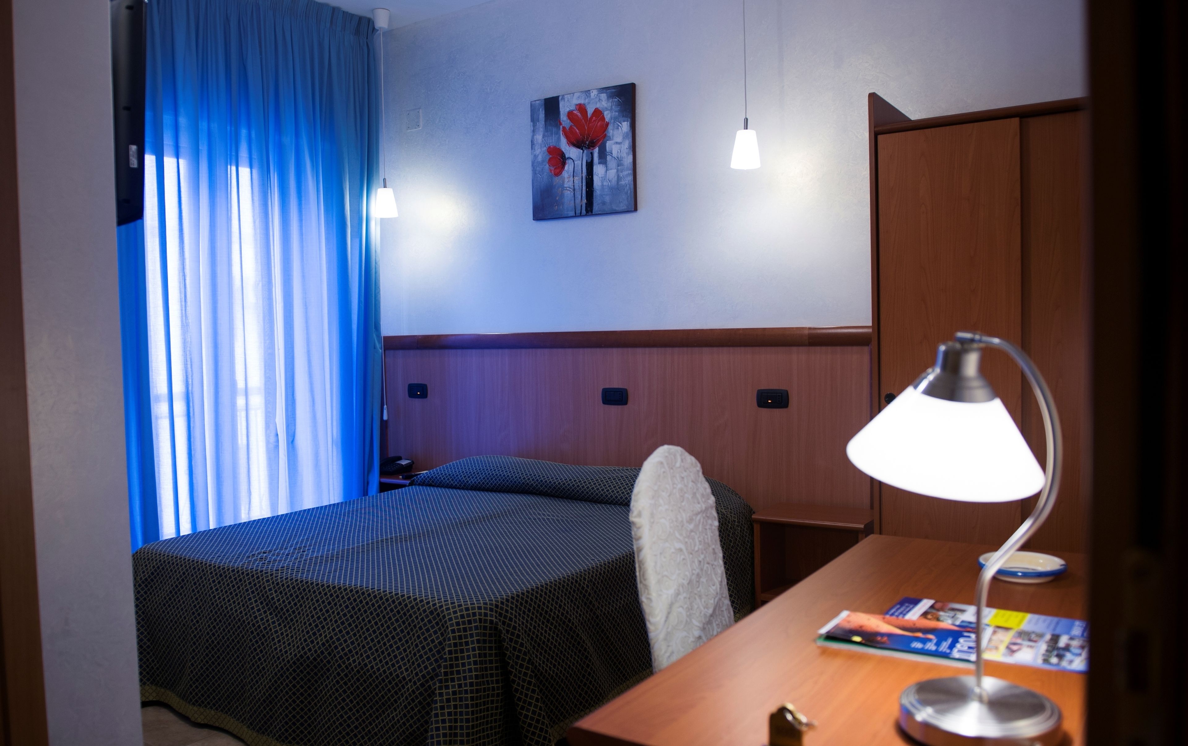 Habitación de hotel con cama, escritorio con lámpara, una silla, un cuadro en la pared y ventana con cortinas azules.