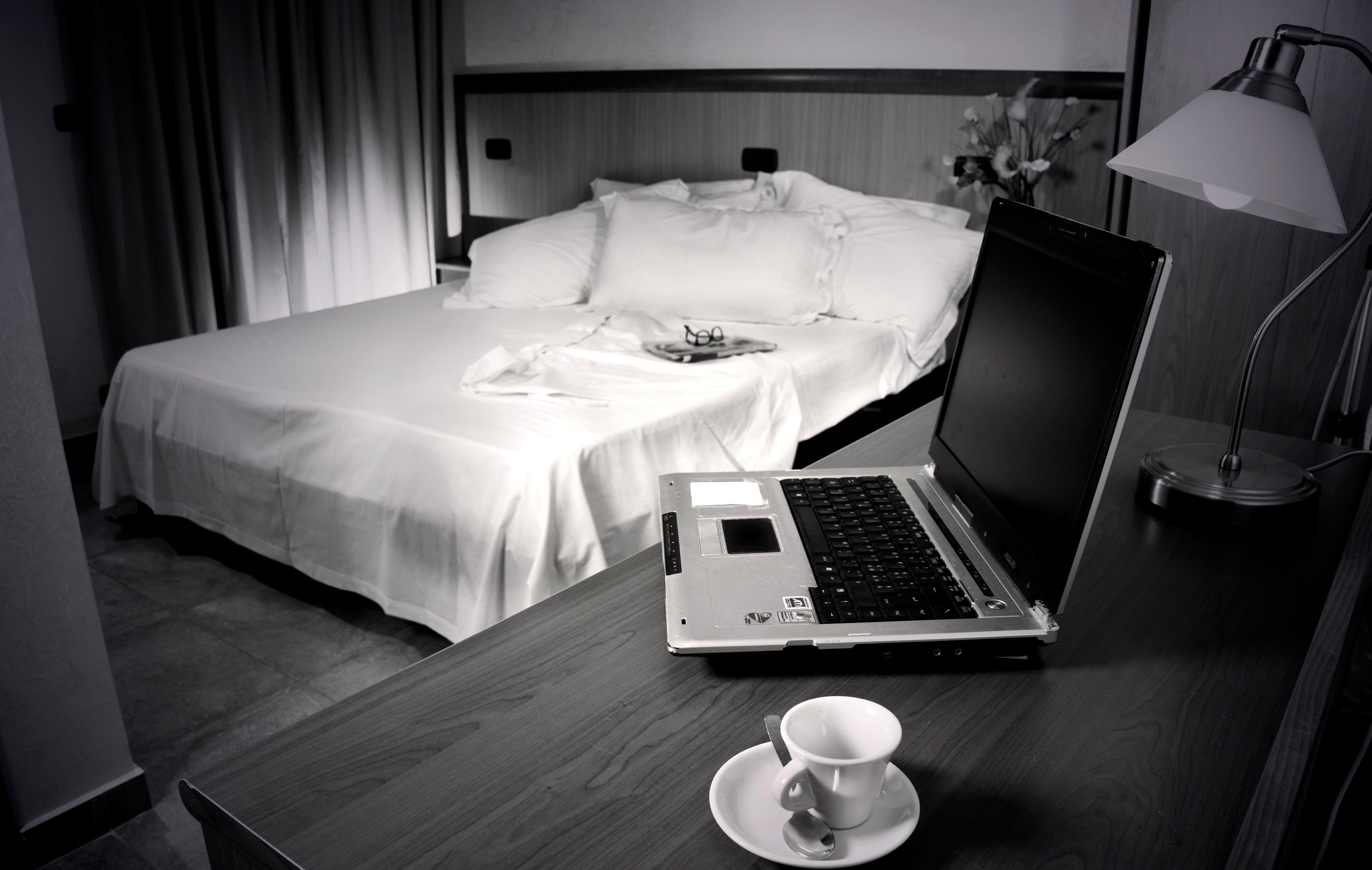 A hotel room with a bed, a laptop on a wooden desk, a cup on a saucer, and a lamp.