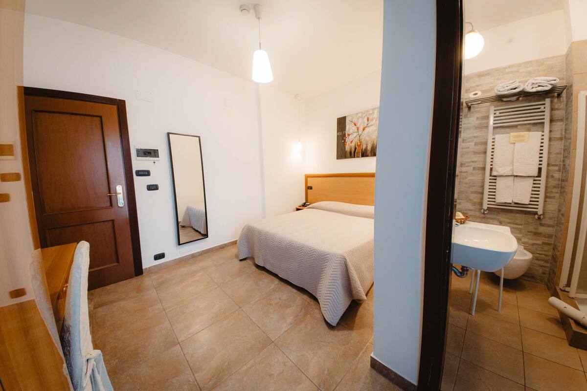 Een slaapkamer met een bed, nachtkastje, spiegel, deur en een badkamer met wastafel en toilet.