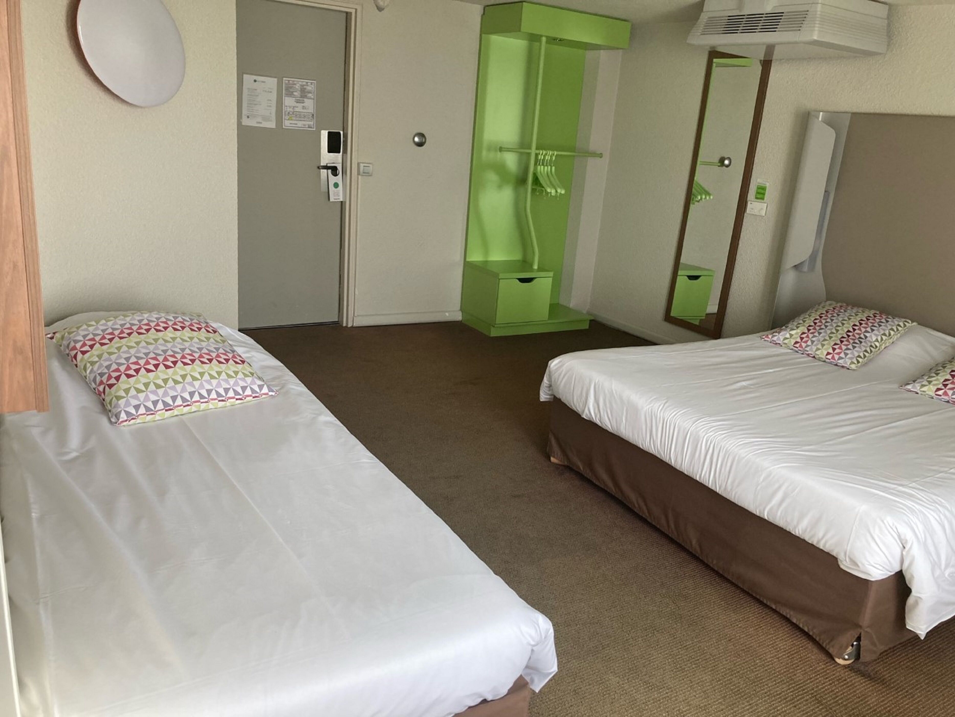 A hotel room with two single beds, a green wardrobe, a mirror, and a door with a lock and keycard reader.