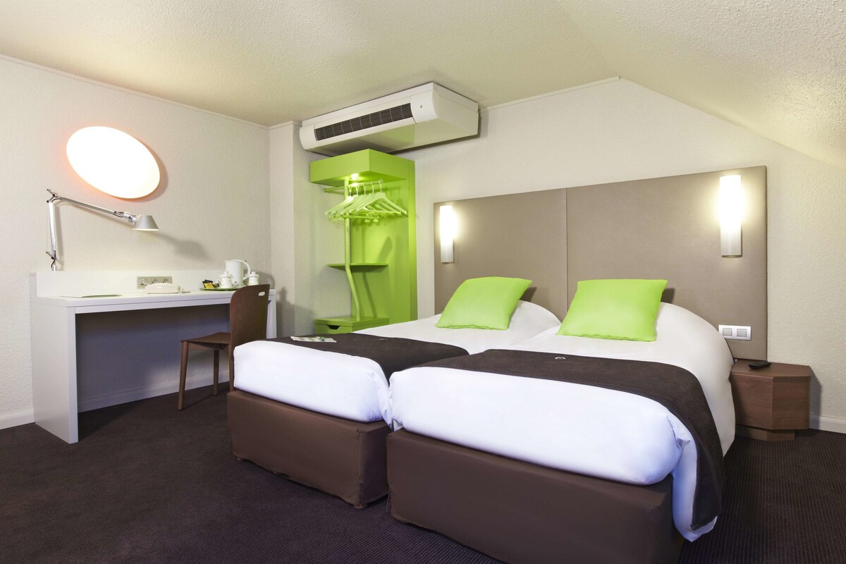Habitación de hotel con escritorio, dos camas con almohadas verdes, una silla, perchero y equipo de aire acondicionado.