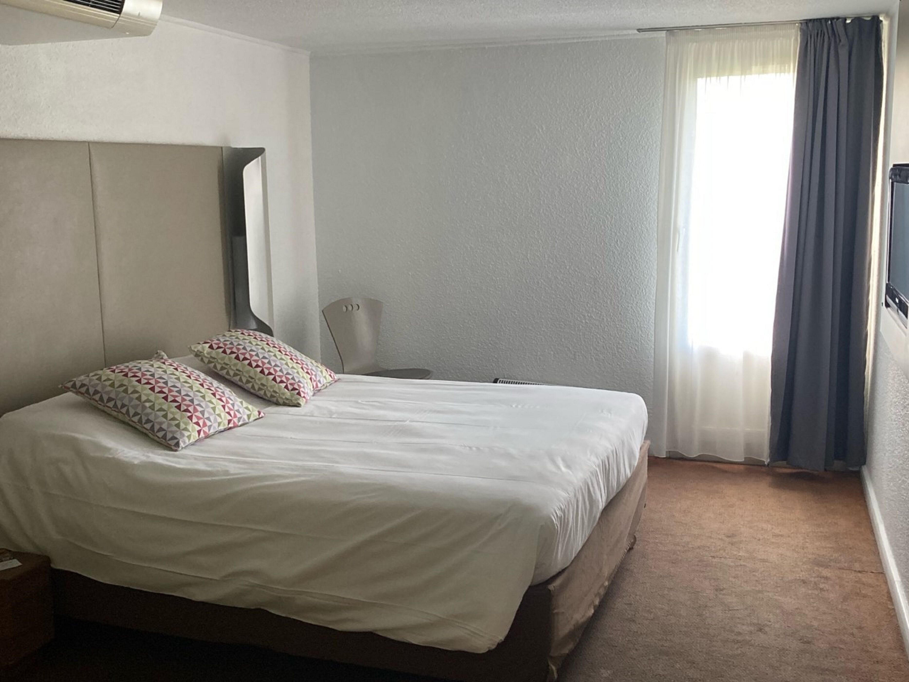 Habitación de hotel con una cama grande, dos almohadones estampados, una silla, un televisor y una ventana con cortinas.