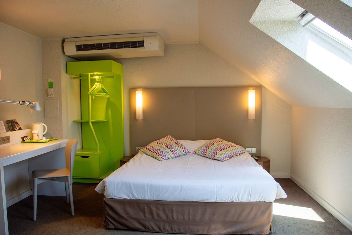 Une chambre d’hôtel avec un lit, un bureau, une chaise, une armoire verte et une fenêtre.