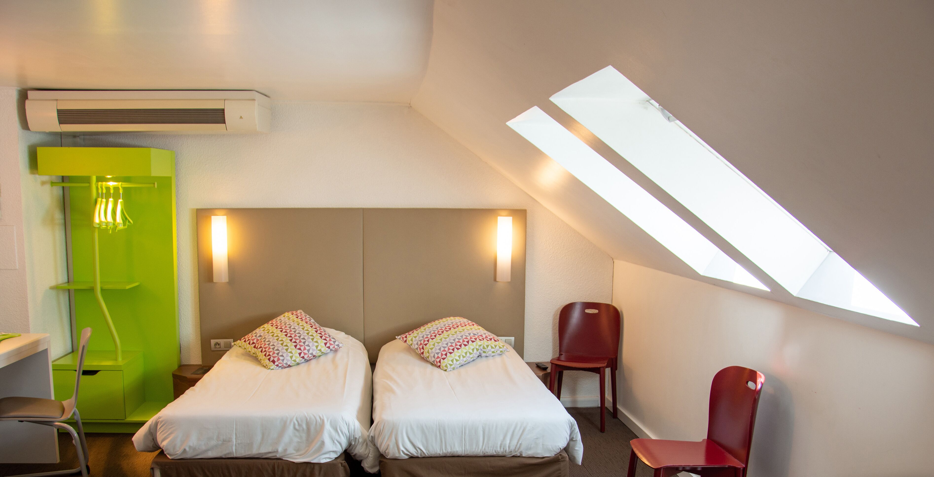 A hotel room with two beds, a desk, a chair, and a skylight.