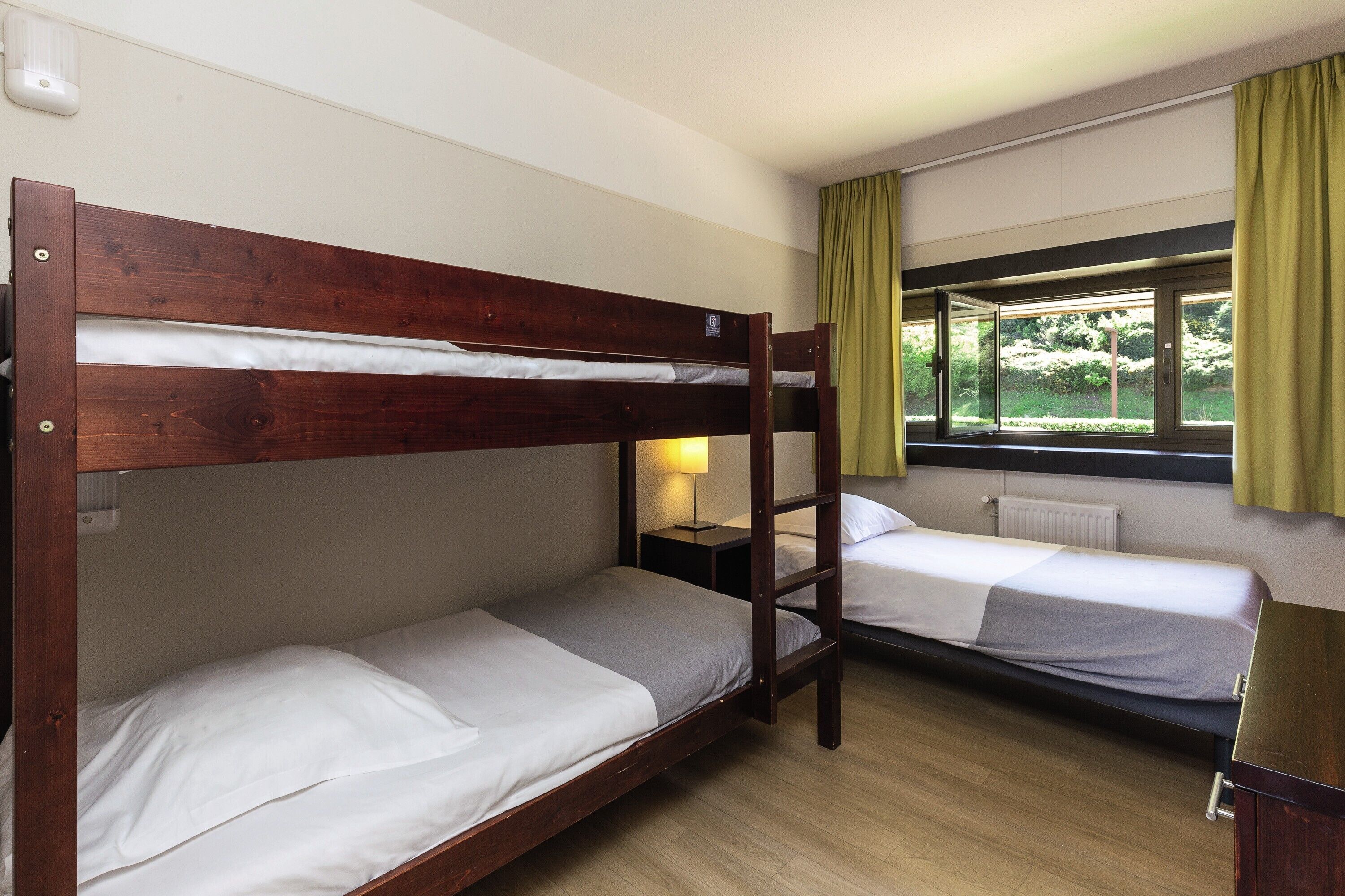 Ein Zimmer mit zwei Etagenbetten, einem Nachttisch mit Lampe, einem Heizkörper und einem Fenster mit Vorhängen.