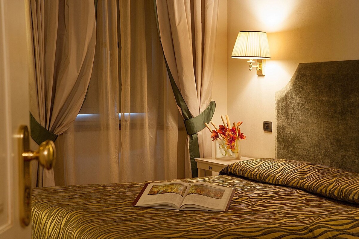 A hotel room with a bed, a bedside table with a lamp, a vase with flowers, and curtains.