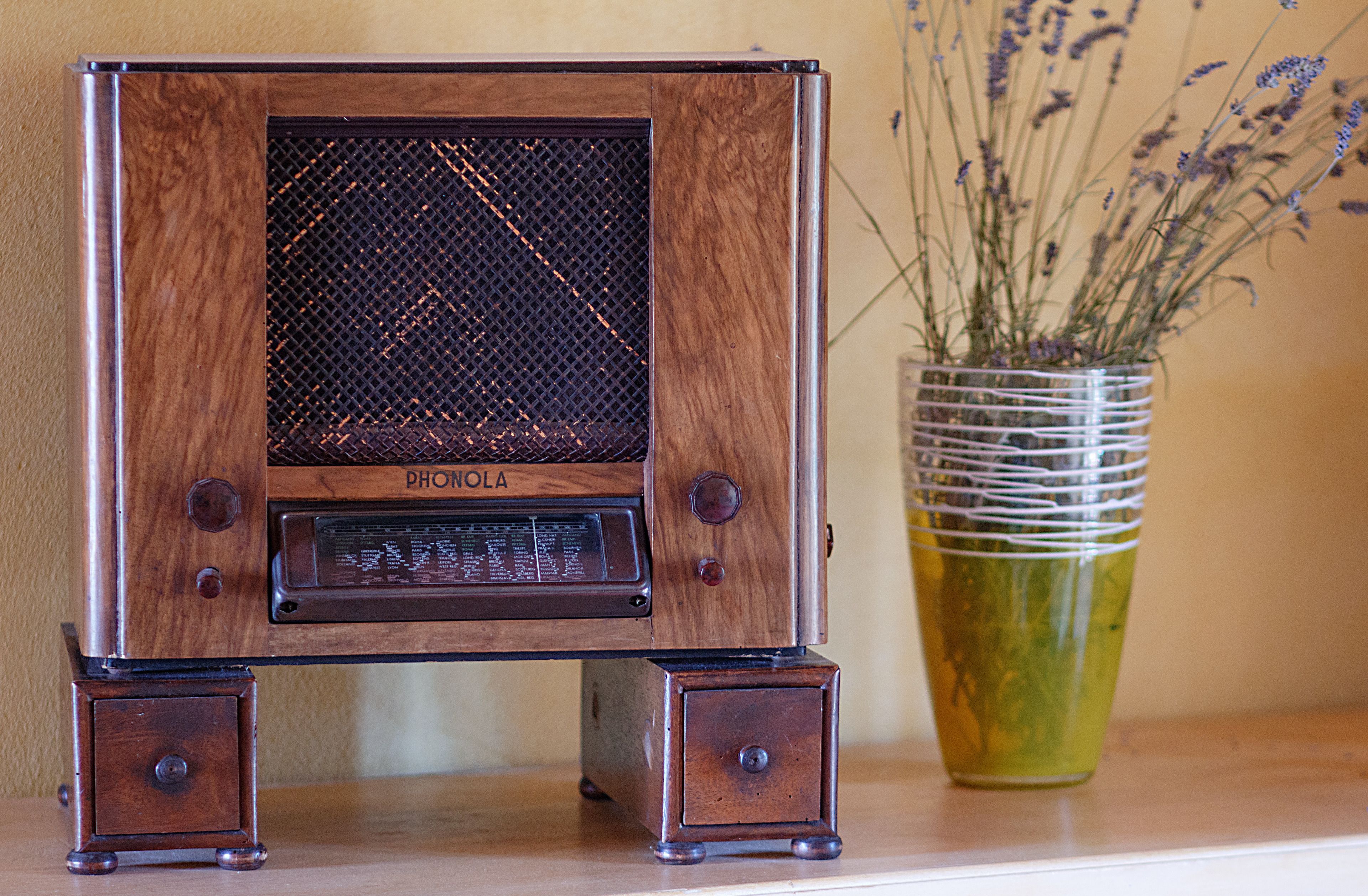 Una radio de madera antigua con rejilla de altavoz de malla y una pantalla pequeña, sobre una superficie de madera junto a un florero de vidrio con lavanda seca.