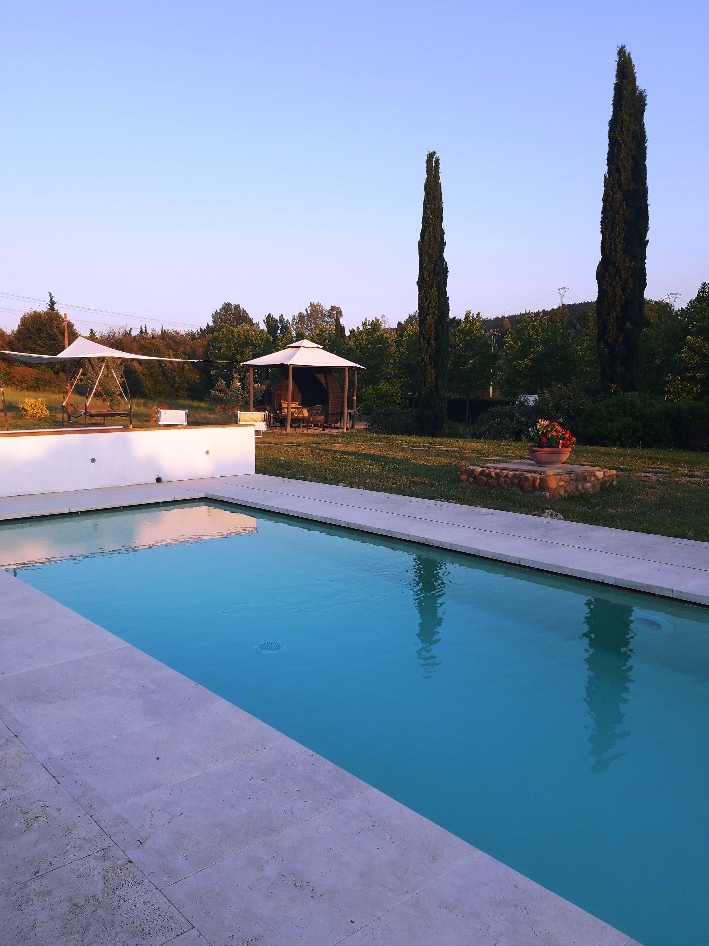Una piscina rettangolare con solarium bianco, area relax coperta e alberi alti sullo sfondo.