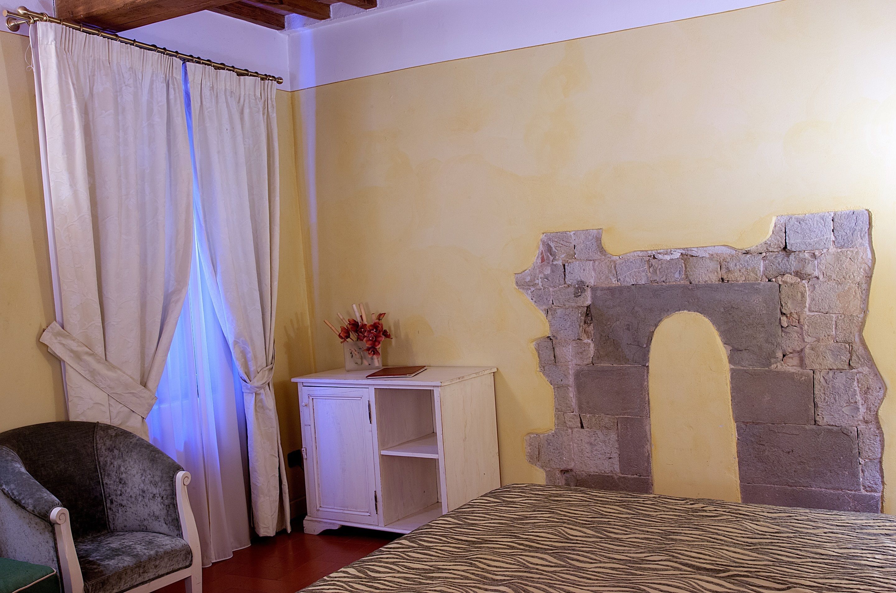 Una stanza con un letto, una sedia, un piccolo armadietto bianco, una tenda e un arco in pietra.