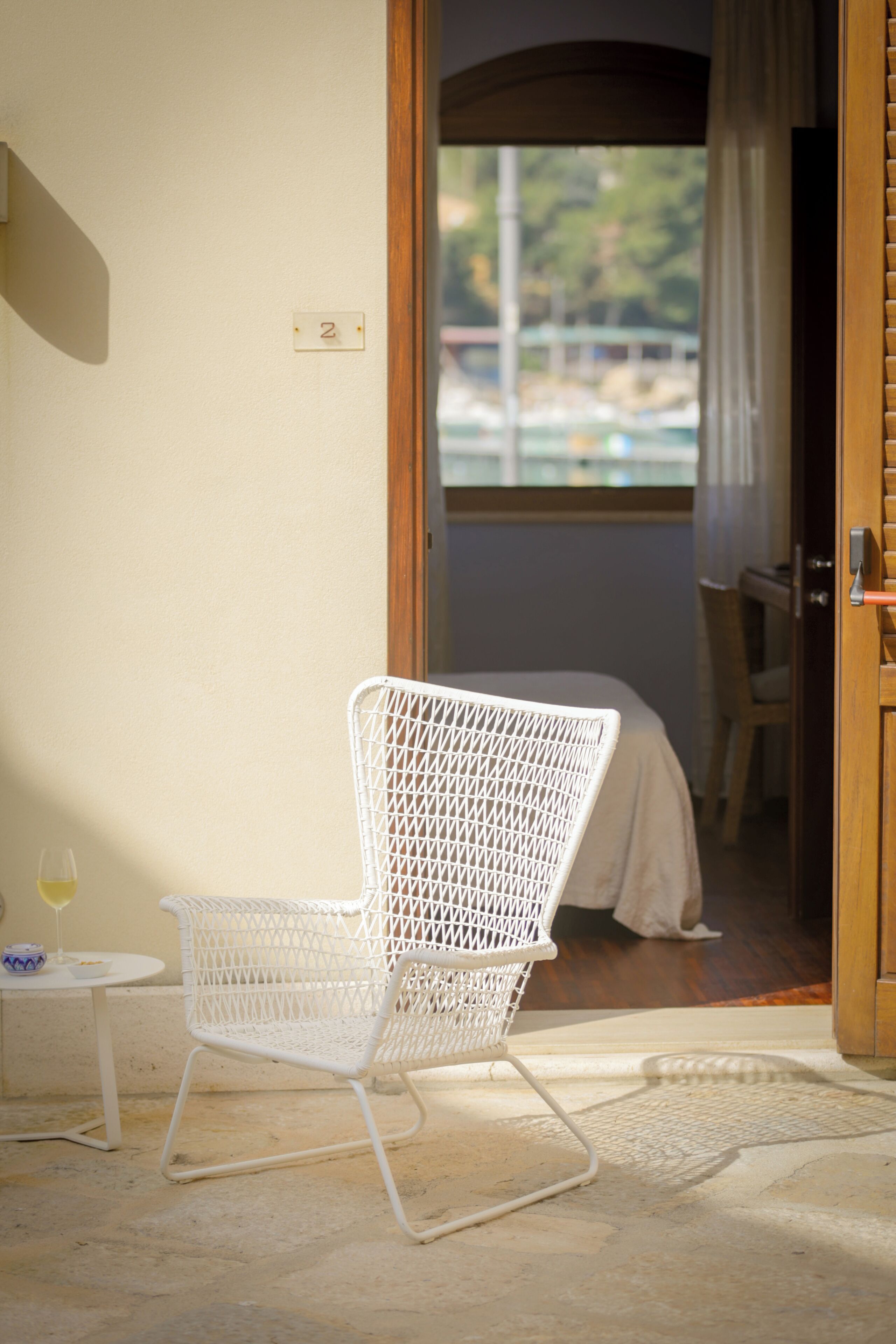 A white wicker chair in a room with a wooden door, a small table with a glass of wine, and a number '2' on the wall.