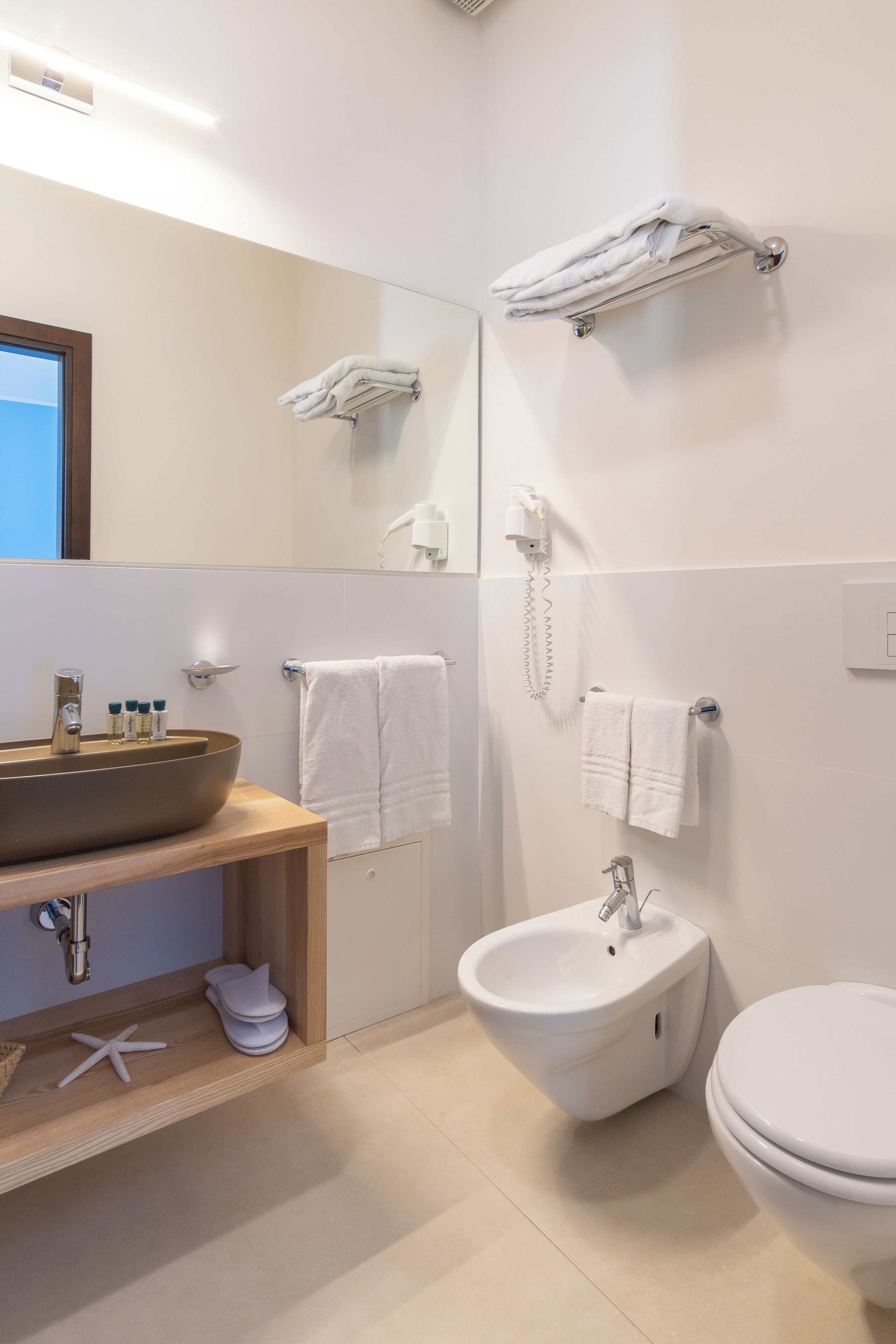 A modern bathroom with a large sink, bidet, and toilet. There are multiple towels and a hairdryer on the wall.