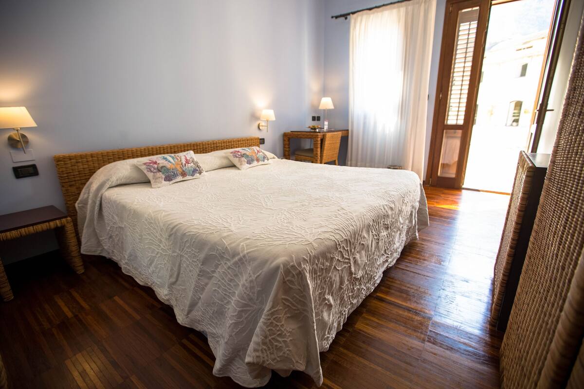 Een slaapkamer met een groot bed, nachtkastjes, een klein bureau en een raam met gordijnen.