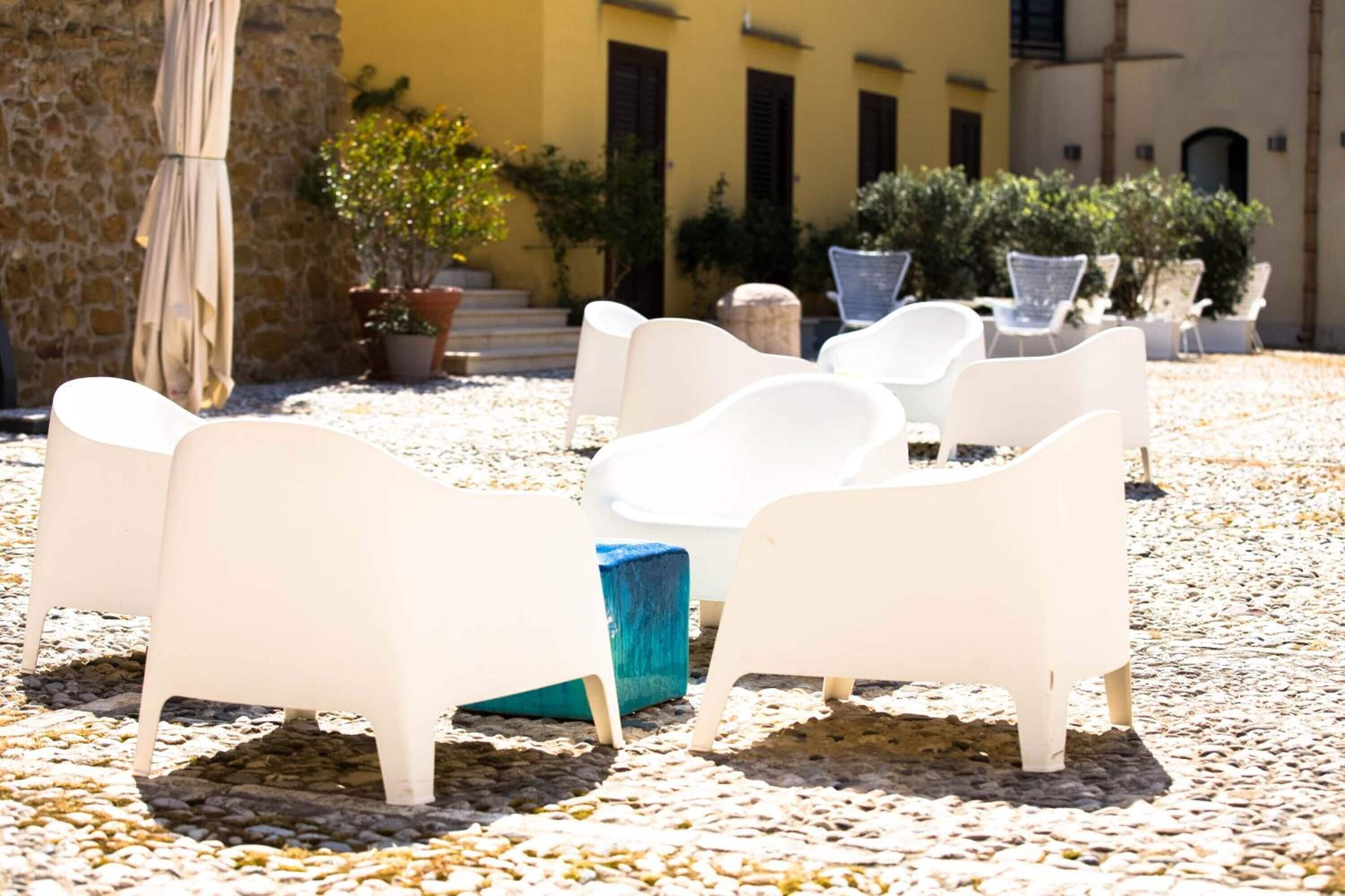 A courtyard with white chairs and a blue ottoman, surrounded by potted plants and a stone wall.