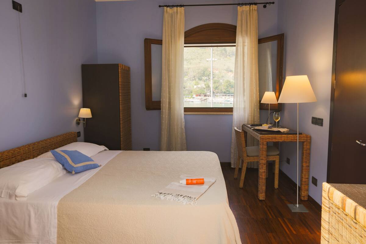 Hotelkamer met een bed, een bureau met lamp, een stoel, een raam met gordijnen en uitzicht op een waterpartij.
