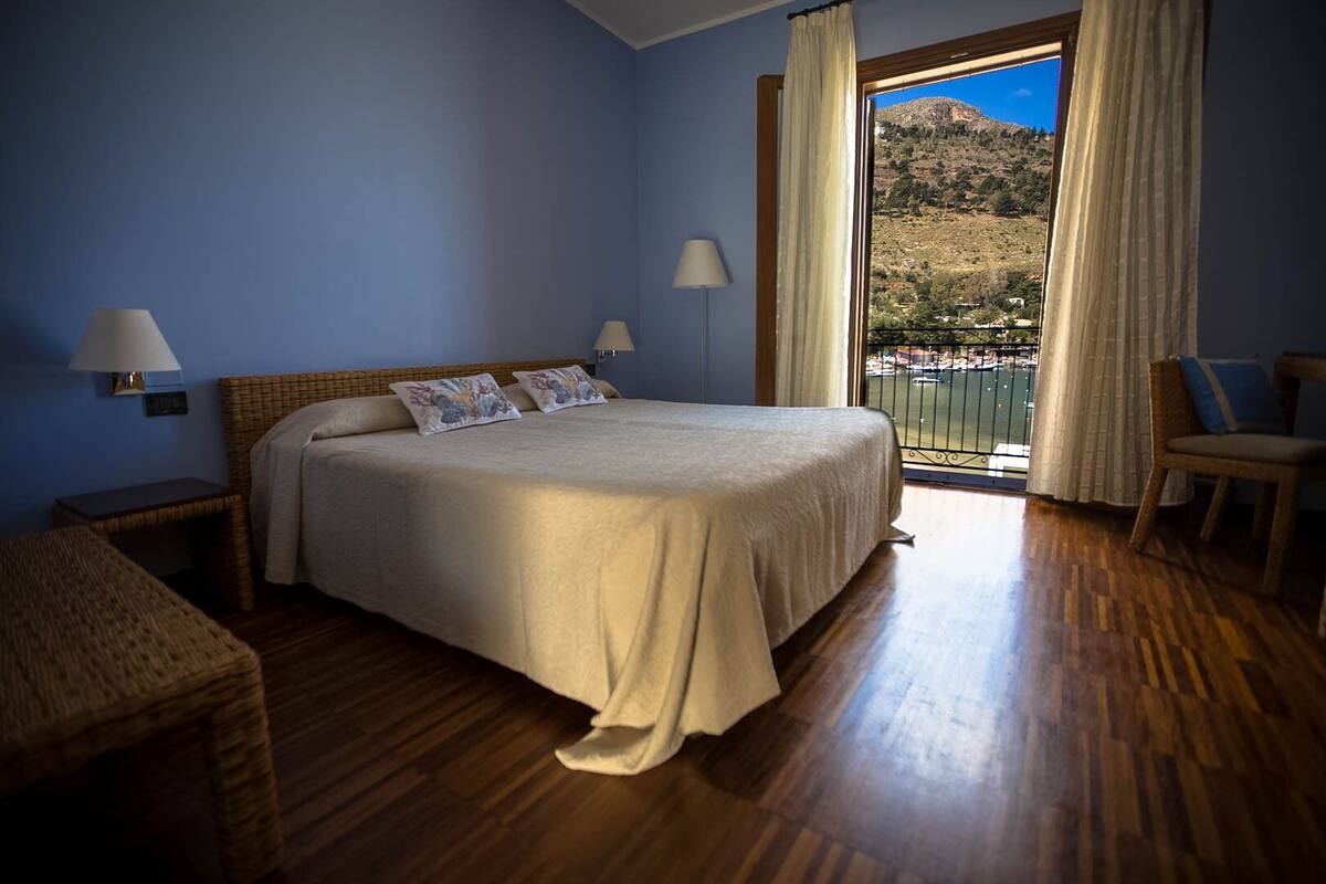 Een slaapkamer met een groot bed, twee bedlampen, een rieten stoel en uitzicht op een bergachtig landschap vanuit een balkon.