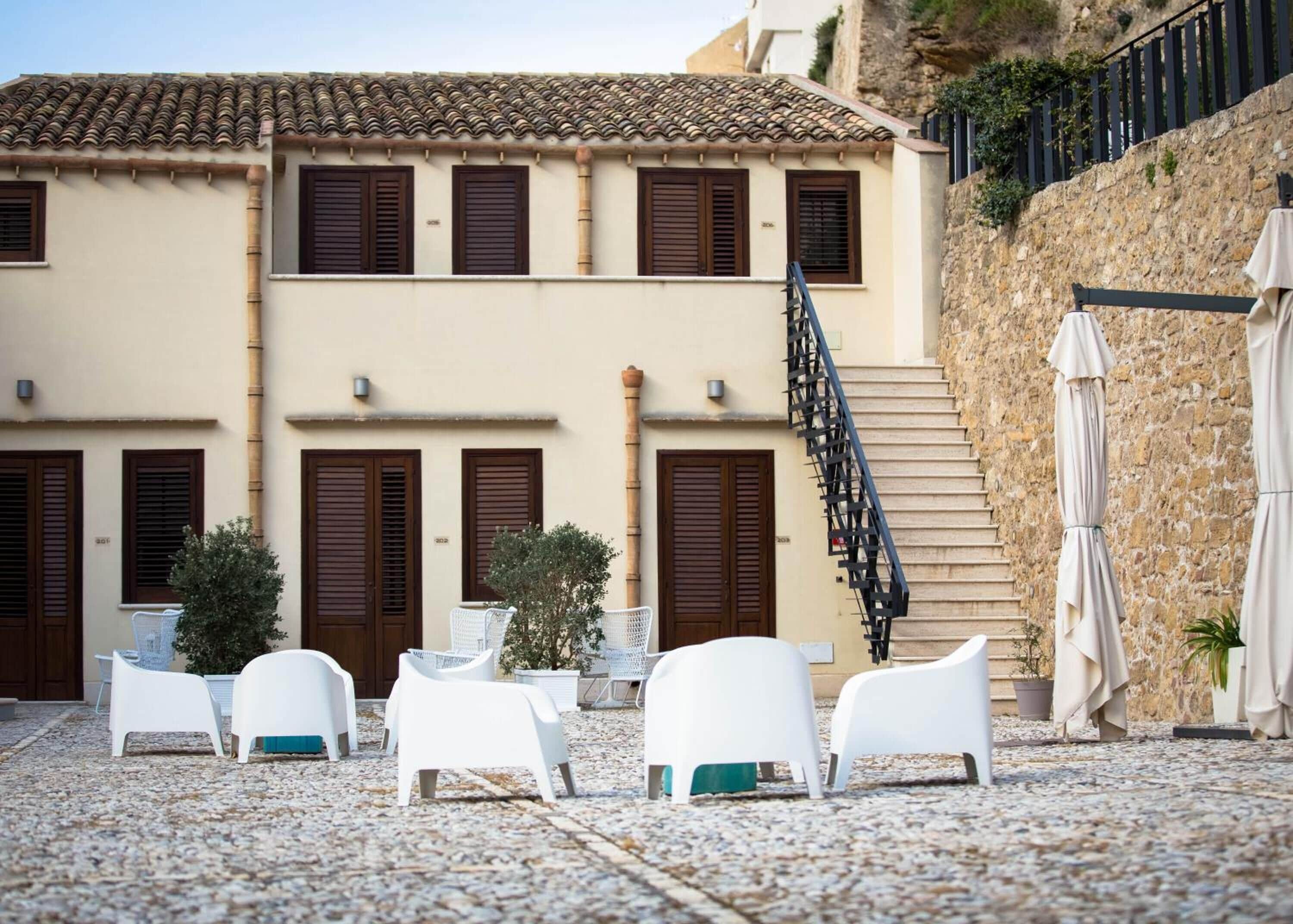 Un bâtiment doté d’un toit en tuiles, de nombreuses fenêtres avec volets, d’un escalier et d’un espace extérieur aménagé avec des parasols.