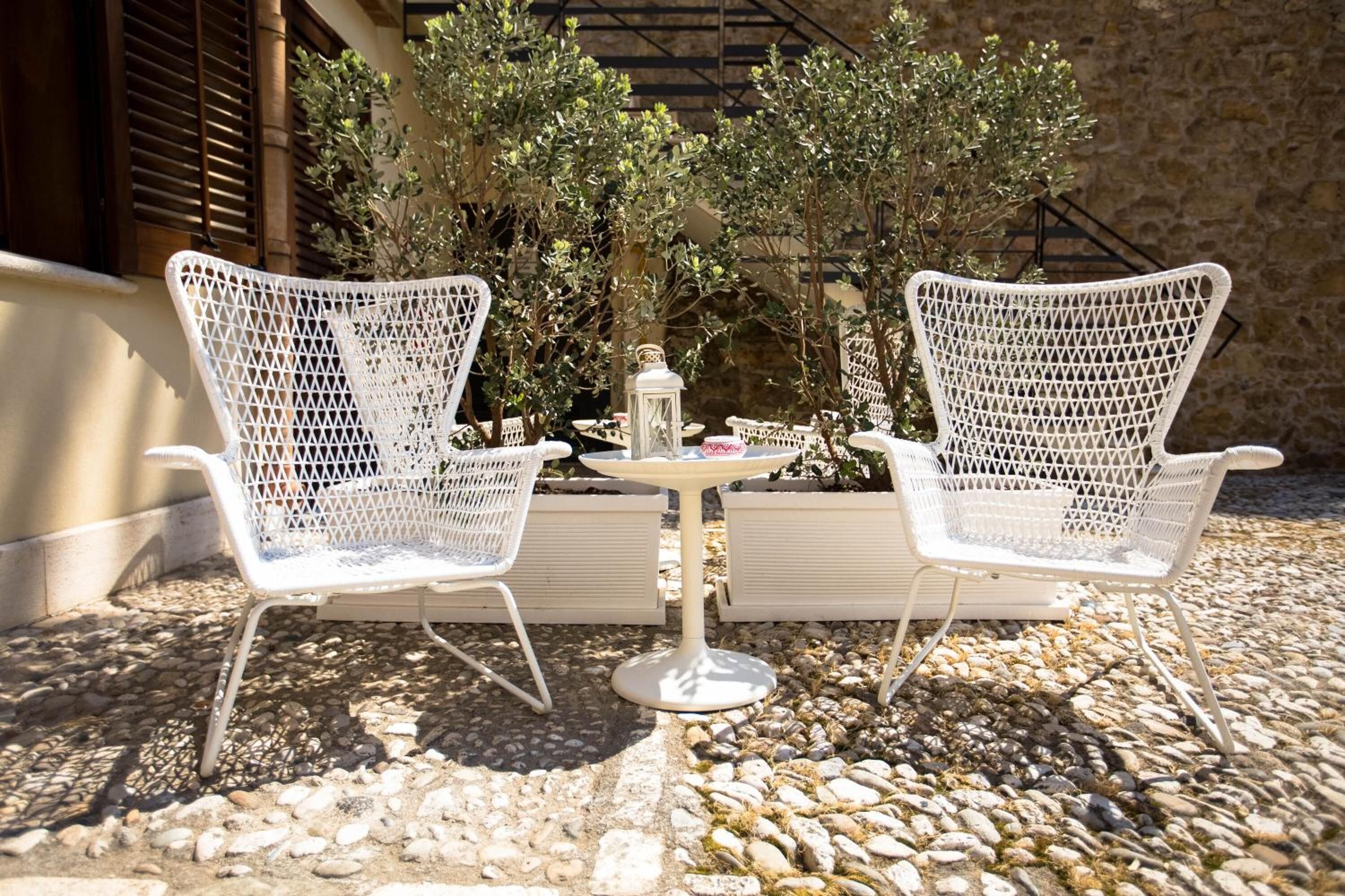 Two white wicker chairs with a small round table between them, set on a gravel surface with plants and a stone wall in the background.
