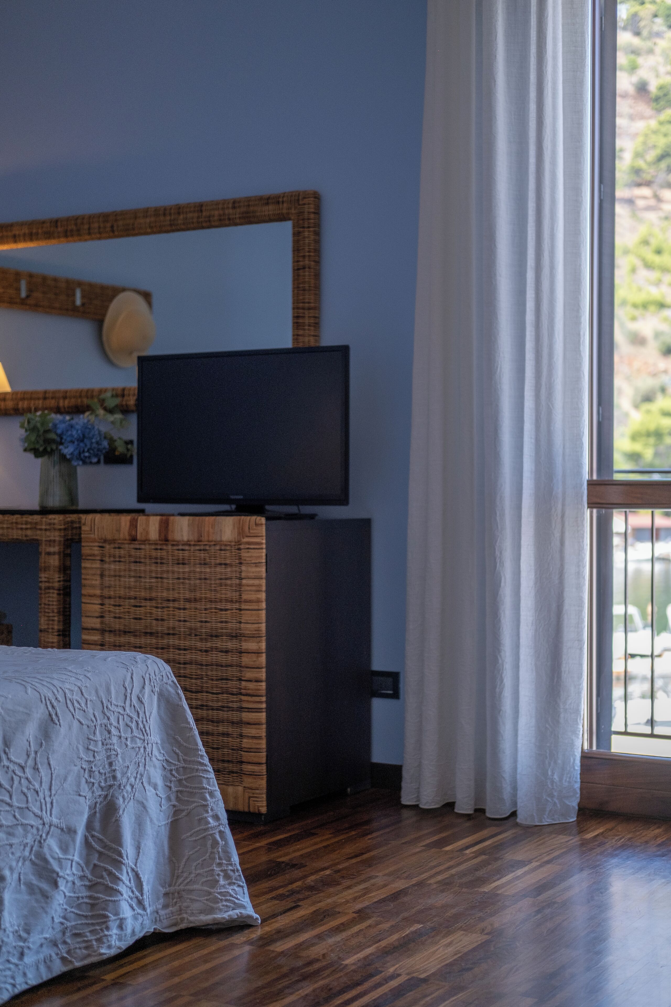 Una habitación con cama, televisión sobre un mueble de madera, ventana con cortinas y vista a zonas verdes.