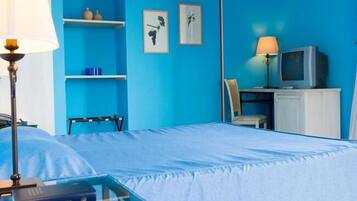 Habitación de hotel con pared azul, cama con colcha blanca, mesita de noche con lámpara, televisor sobre un mueble y cuadros enmarcados en la pared.