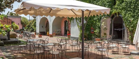 An outdoor patio with tables and chairs, covered by a large umbrella, surrounded by greenery and architectural arches.