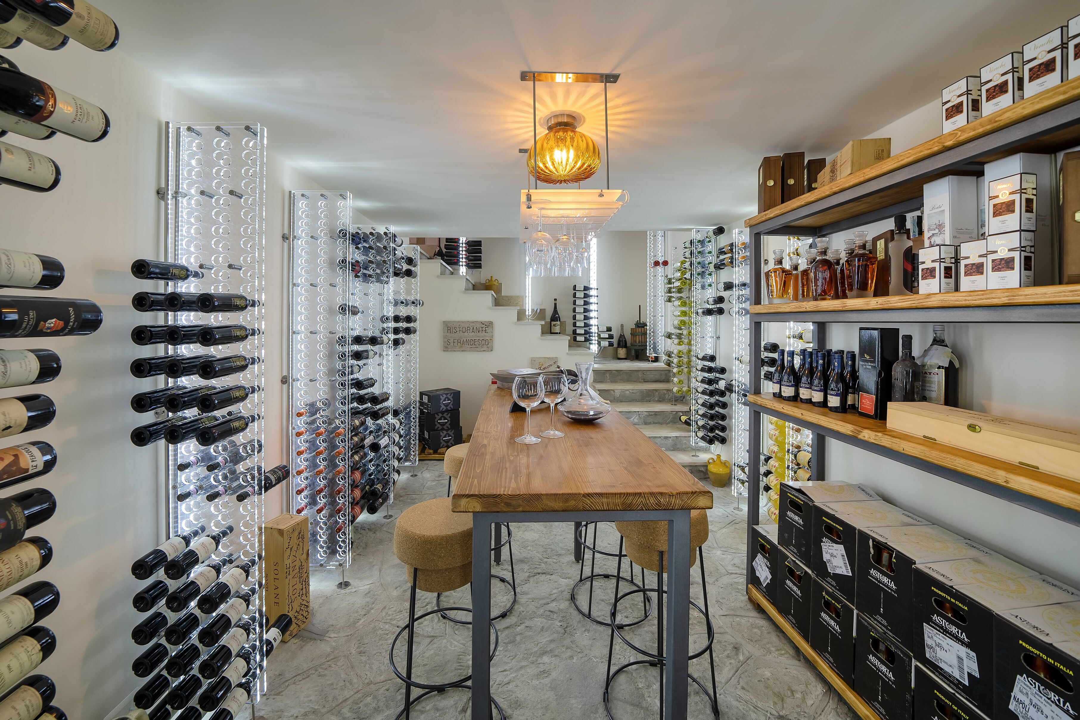 A wine cellar with rows of wine bottles, a wooden table with glasses, and a staircase in the background.