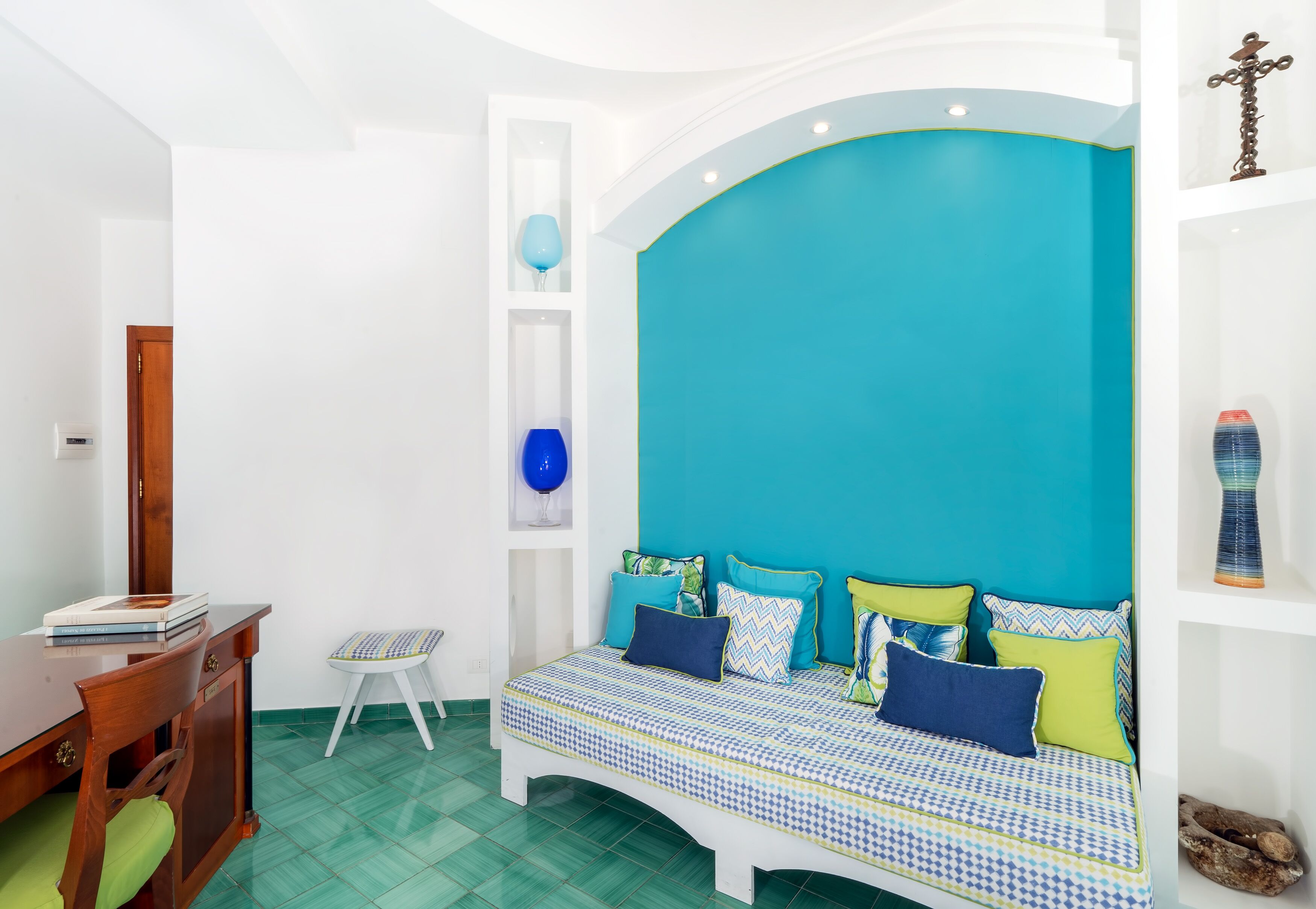 Une chambre avec un mur d’accent de couleur turquoise, un lit avec une literie à motifs, un bureau en bois et un petit tabouret.