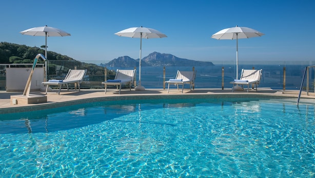 A pool with clear blue water, surrounded by white lounge chairs and umbrellas, with a mountain view in the background.