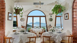 A dining area with tables set for four, large arched windows, hanging plants, and framed pictures on the walls.