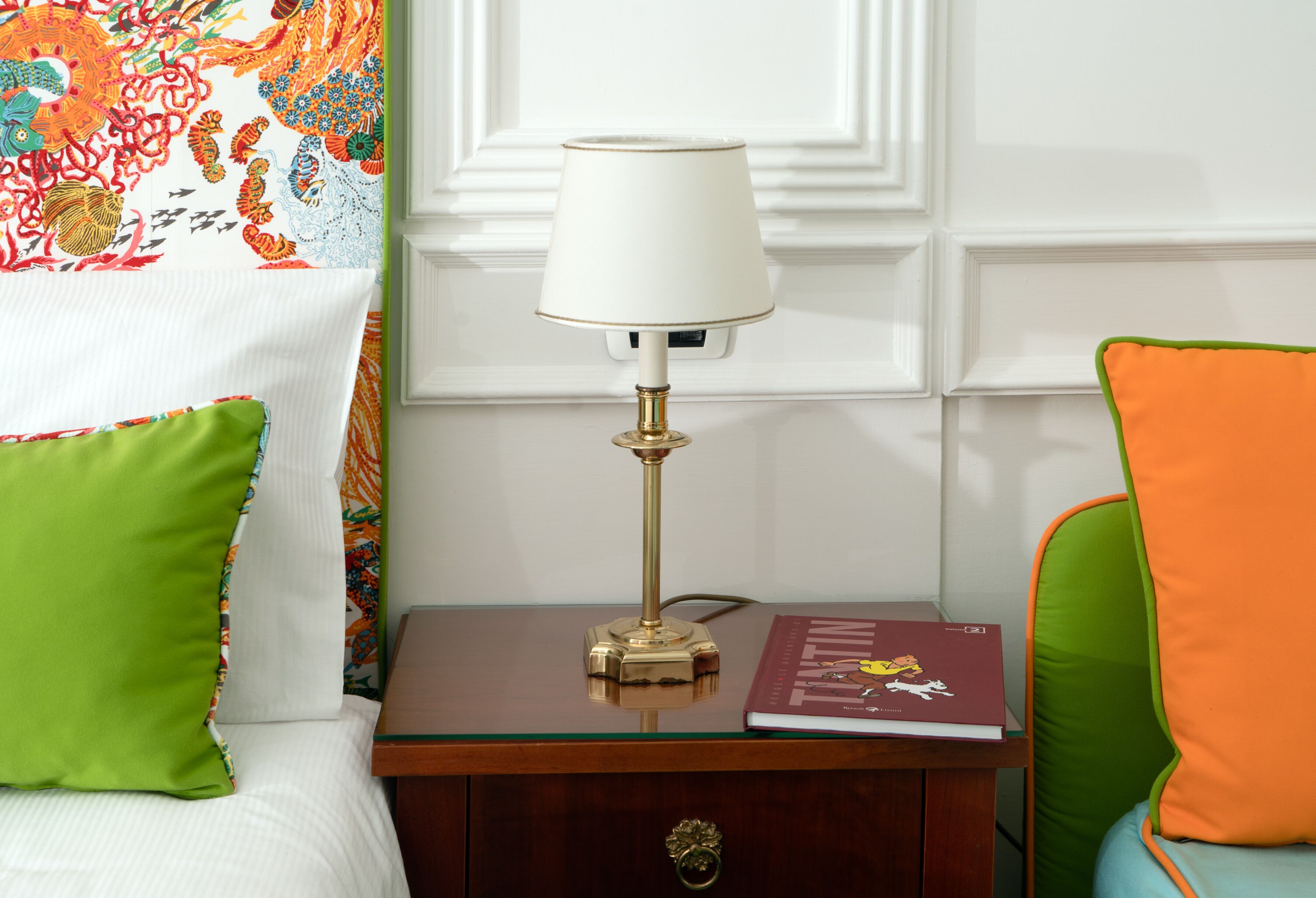 Una cama bien hecha con un almohábil verde, una mesita de noche con una lámpara, un libro y una silla con un cojín naranja.