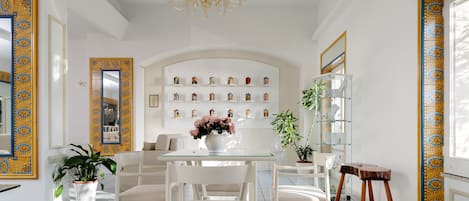 Un comedor moderno con mesa de vidrio, sillas blancas, una araña y una pared con espejos decorativos y estanterías.
