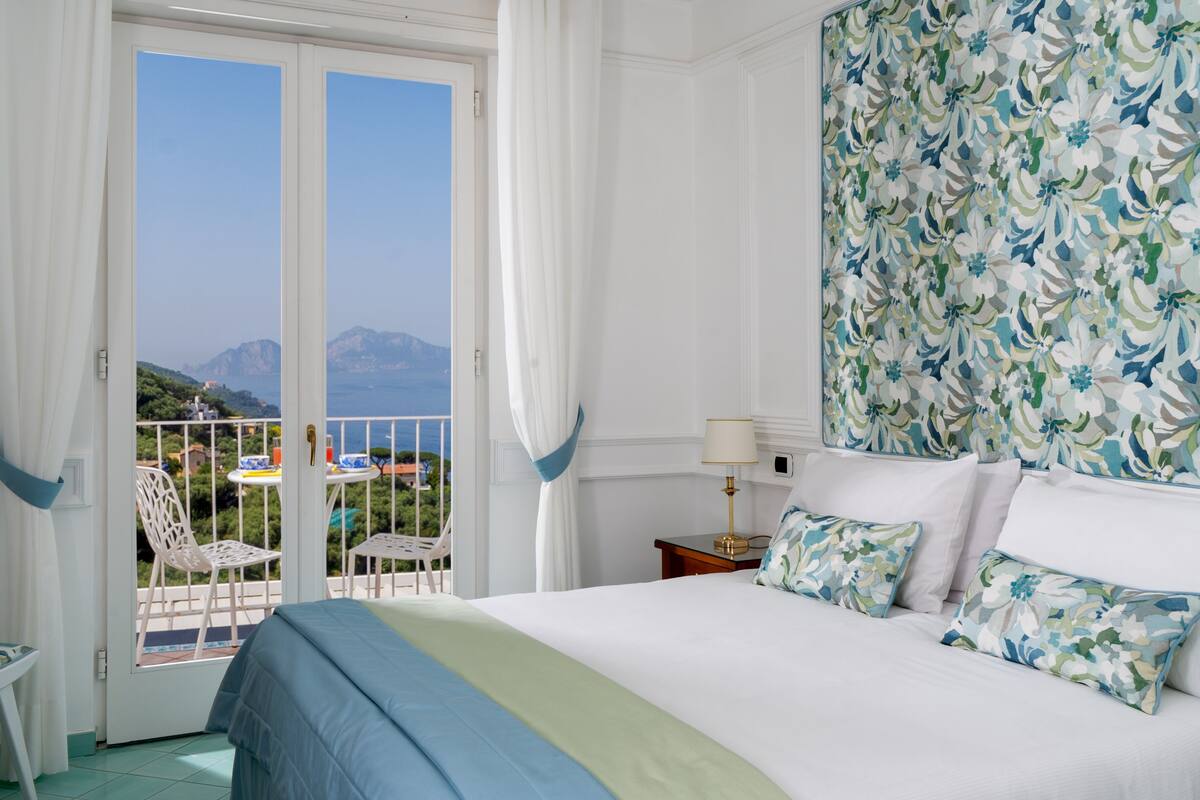 Une chambre d’hôtel avec balcon, un lit agrémenté d’une literie à motifs floraux et une vue sur la mer et les montagnes.