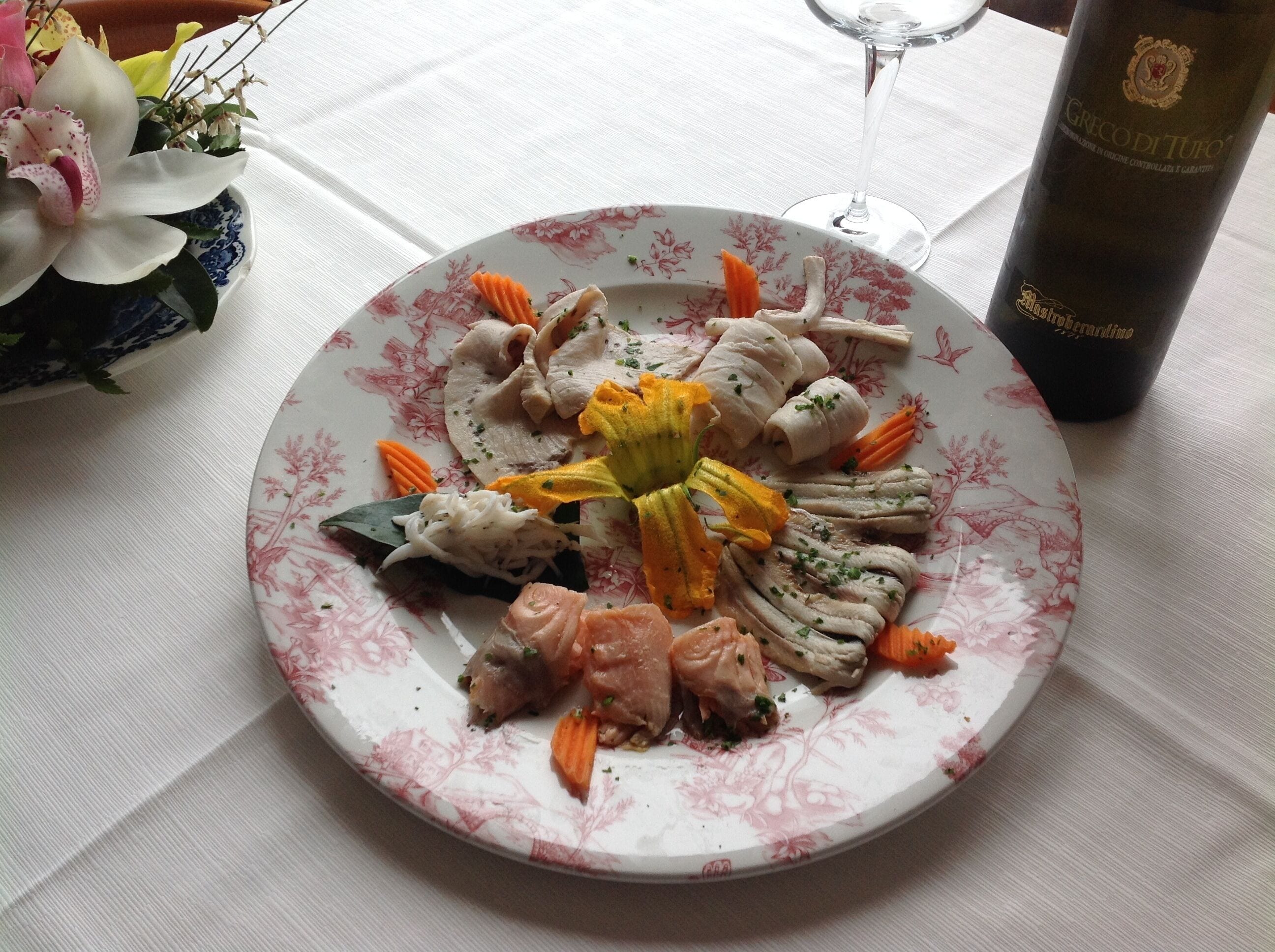 Um prato de comida com vários legumes e carne, um copo de vinho e um vaso decorativo com flores.