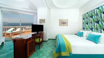 A hotel room with a bed, a desk with a chair, a television, and a large window offering a view of the sea.