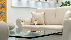 A modern living room with a beige sofa, a glass coffee table with a plate of food and two glasses of wine, and a wall with framed artwork.