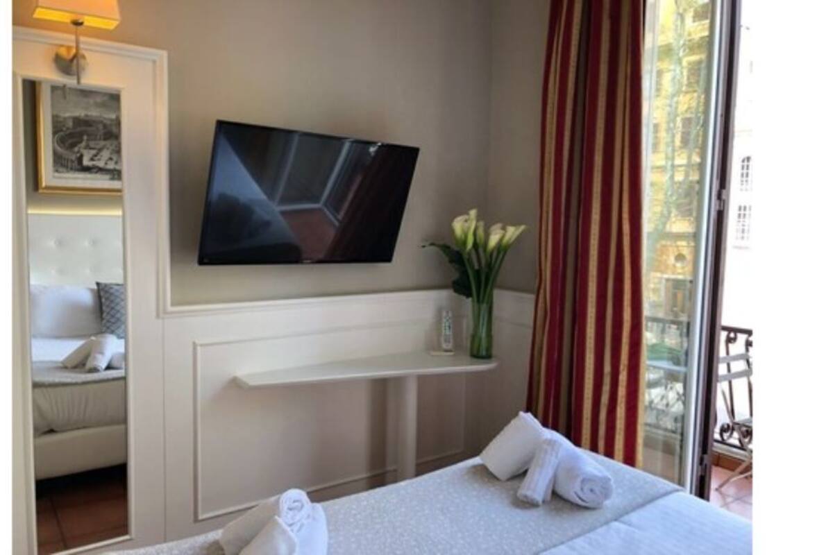 Una habitación de hotel con una cama, un televisor montado en la pared, una mesita con un florero y una ventana con cortinas.