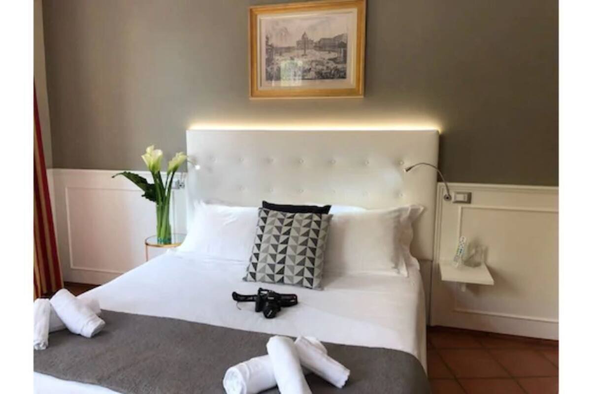 Una cama bien hecha con ropa de cama blanca, una almohada de estampado geométrico y un pequeño jarrón con flores.