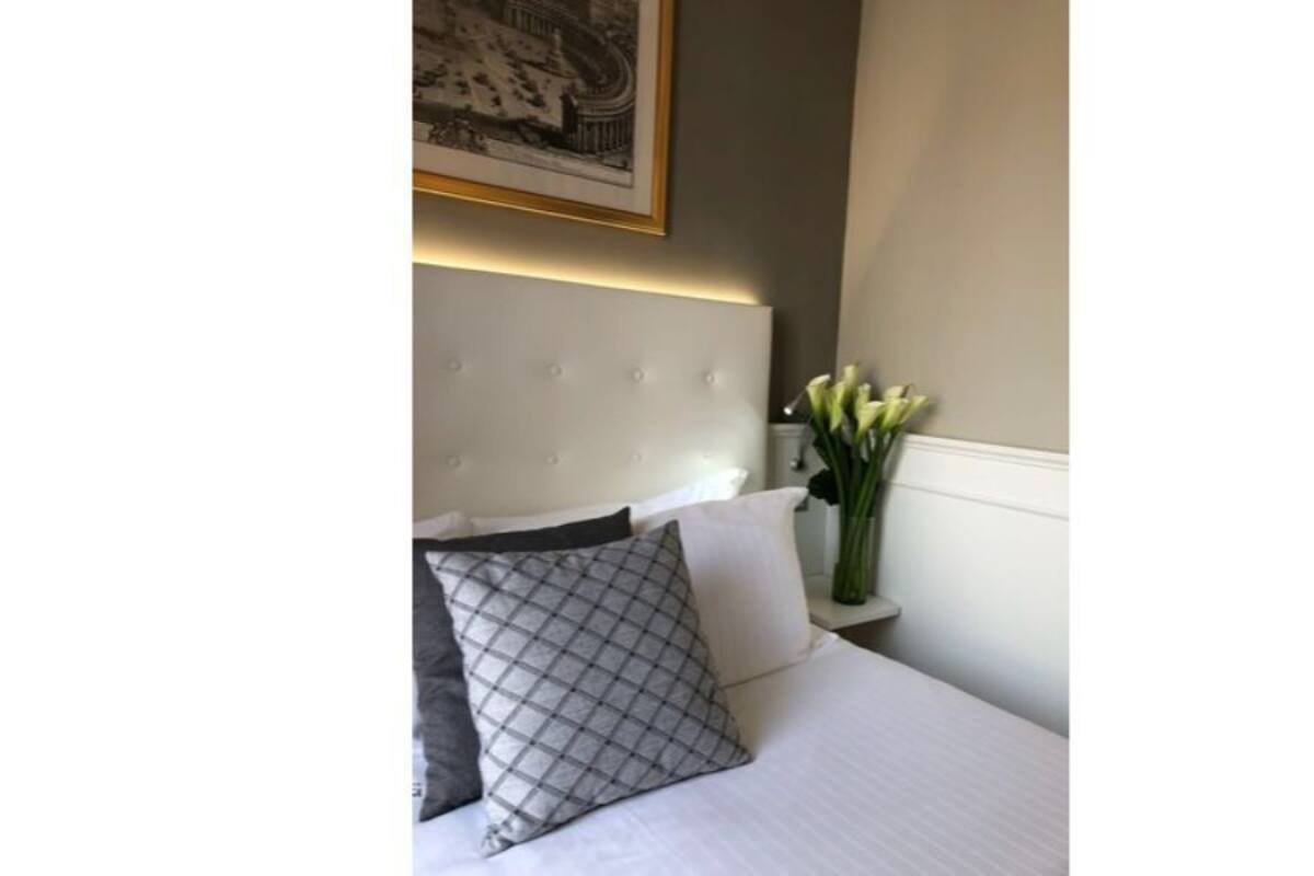 Habitación de hotel con una cama blanca, un almohadón gris y un florero con flores blancas sobre una mesita de noche.