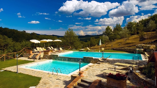 A rectangular pool with a waterfall feature, surrounded by lounge chairs and umbrellas, set against a backdrop of hills and a clear sky.