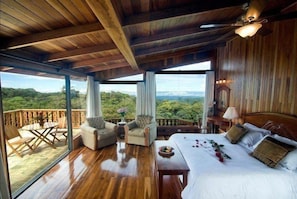 A wooden-floored room with a large bed, a balcony with wicker furniture, and a view of the outdoors. - Belmar (Monteverde)