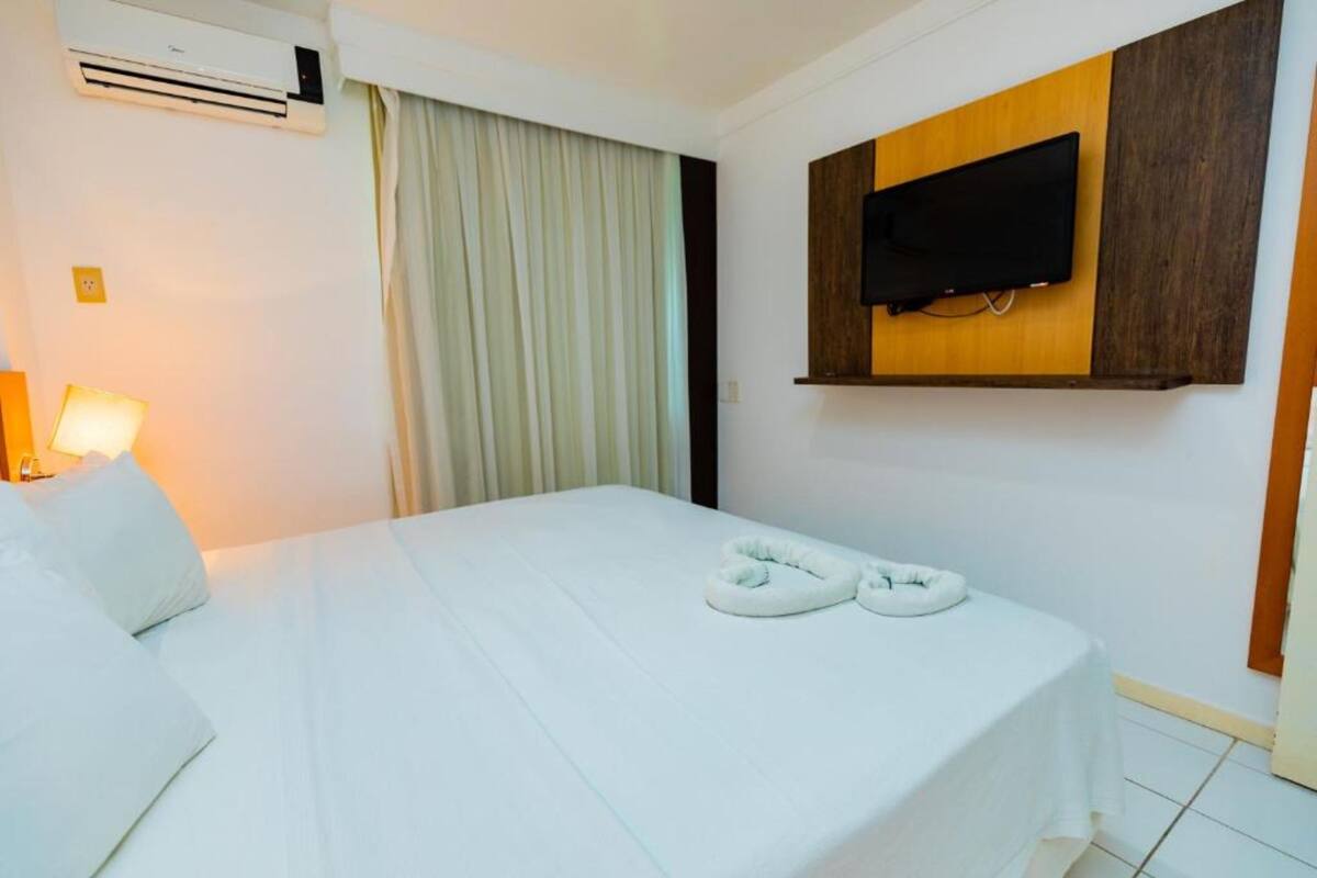 A hotel room with a bed, a TV mounted on the wall, and an air conditioning unit.