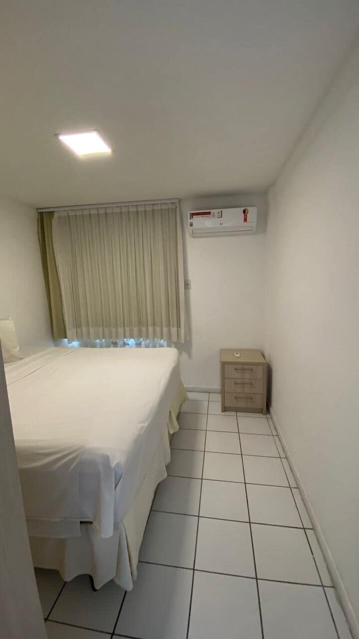 A hotel room with a bed, a window with curtains, a tiled floor, a wall-mounted air conditioning unit, and a small wooden cabinet.