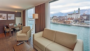 A hotel room with a large window offering a view of a city and a river. There is a sofa, a dining table with chairs, and a lamp. - H4 Hotel Solothurn (Solothurn)