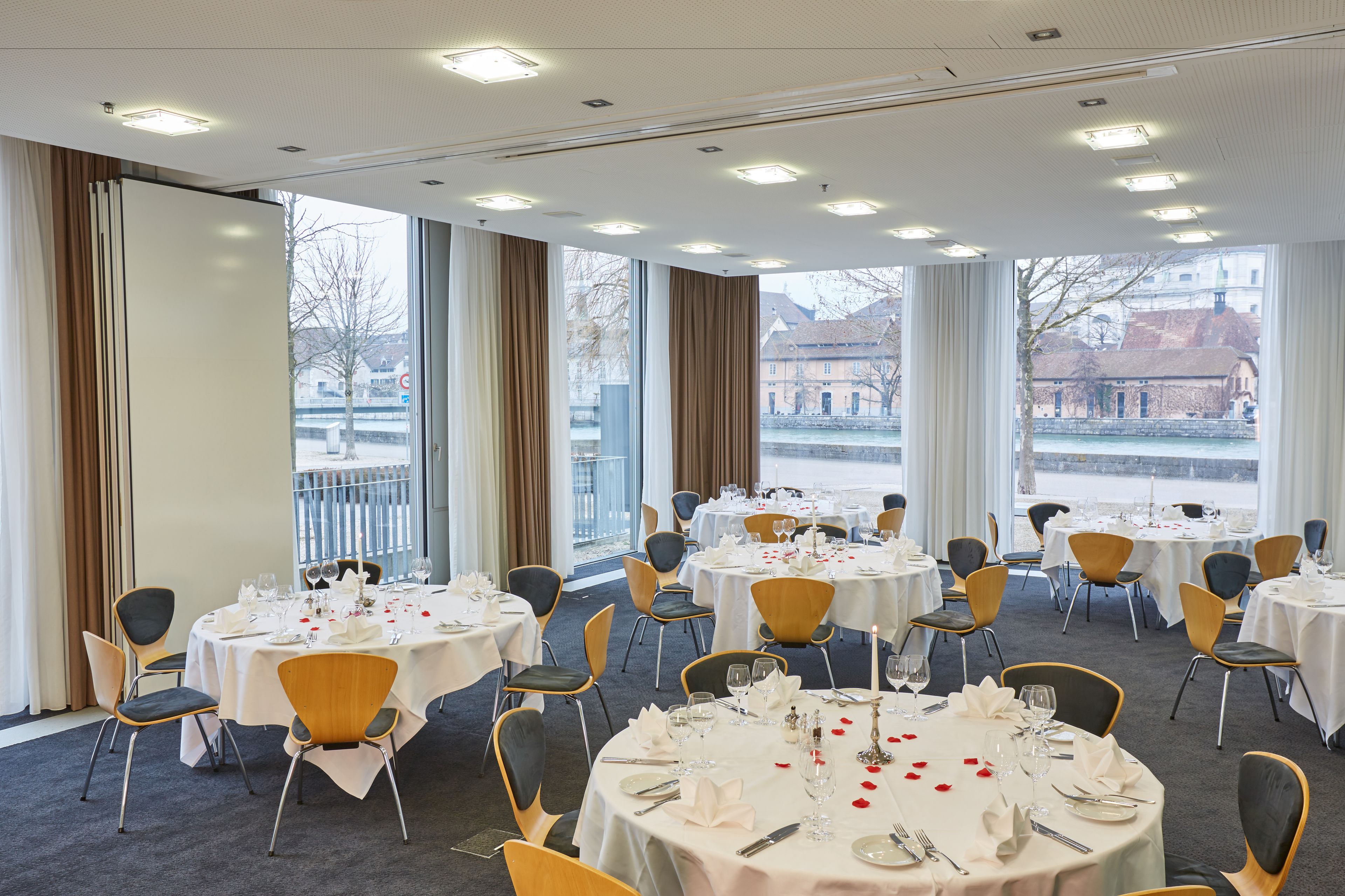 A hotel dining area with round tables set for a meal, large windows with curtains, and a view of buildings and a body of water.