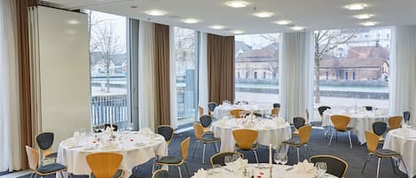A hotel dining area with round tables set for a meal, large windows with curtains, and a view of buildings and a body of water.