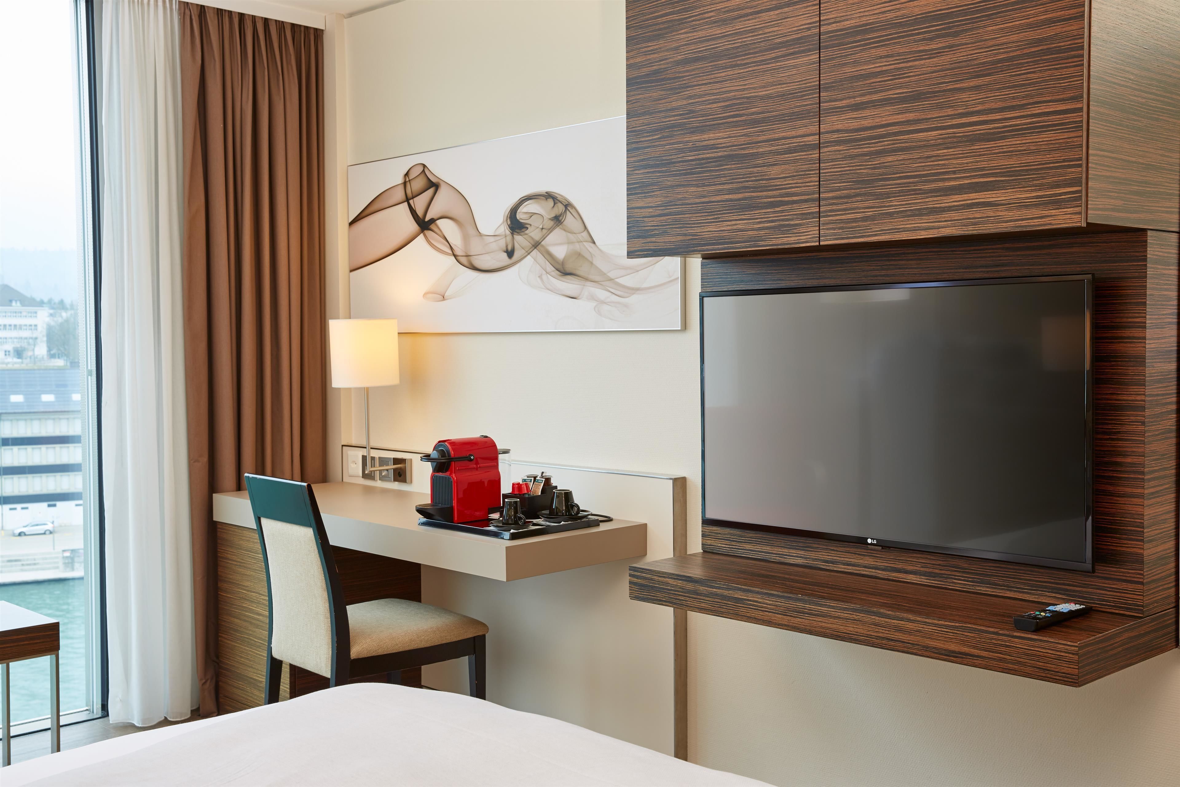 Habitación de hotel con cama, escritorio con cafetera, televisor montado en la pared y ventana con cortinas.