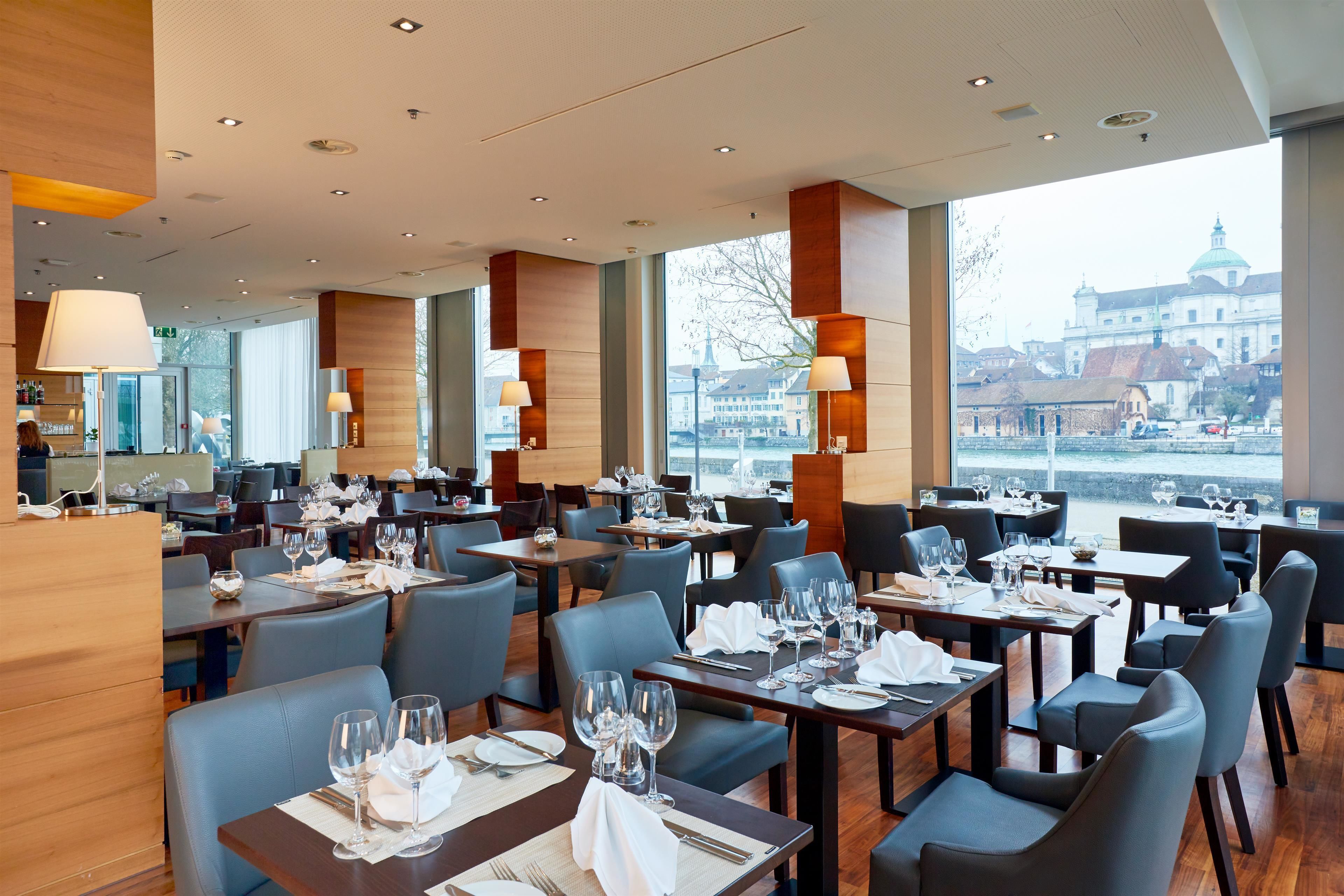 A modern restaurant with multiple dining tables, chairs, and large windows offering a view of a historic building.